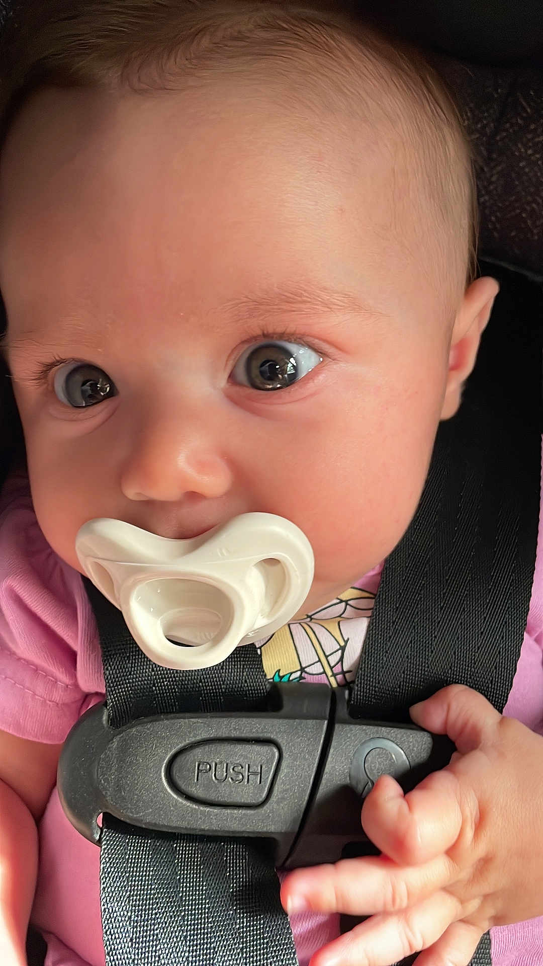 Lisboa is registered to the contest to win money with this photo: baby, infant, pacifier, car_seat, child, face, hand, clothing, pink, closeup, young_child, sitting, seatbelt, cute, curious, portrait, indoor, human, person, newborn