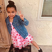 Arianna is registered to the contest to win money with this photo: child, girl, peace_sign, denim_jacket, pink_dress, hearts, welcome_mat, door, window, concrete_floor, black_boots, outdoor, posing, smiling, young, casual_clothing, playful, wall, youth, happy