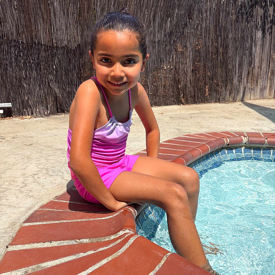 Arianna is registered to the contest to win money with this photo: child, girl, swimsuit, pool, water, brick, outdoor, sunlight, smiling, sitting, playful, summer, young, fence, feet, recreation, leisure, happy, daytime, skin