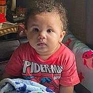 Ke-Shawn is registered to the contest to win money with this photo: blanket, casual_clothing, child, colorful, couch, curious, curly_hair, expression, face, furniture, home, indoor, person, red_shirt, sitting, soft_light, spiderman, toddler, toy, young_child