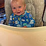baby, child, high_chair, onesie, blue_clothing, smiling, face, indoor, messy_tray, milk_carton_pattern, donut_pattern, cute, infant, happy, person, furniture, wooden_chair, table, home, portrait