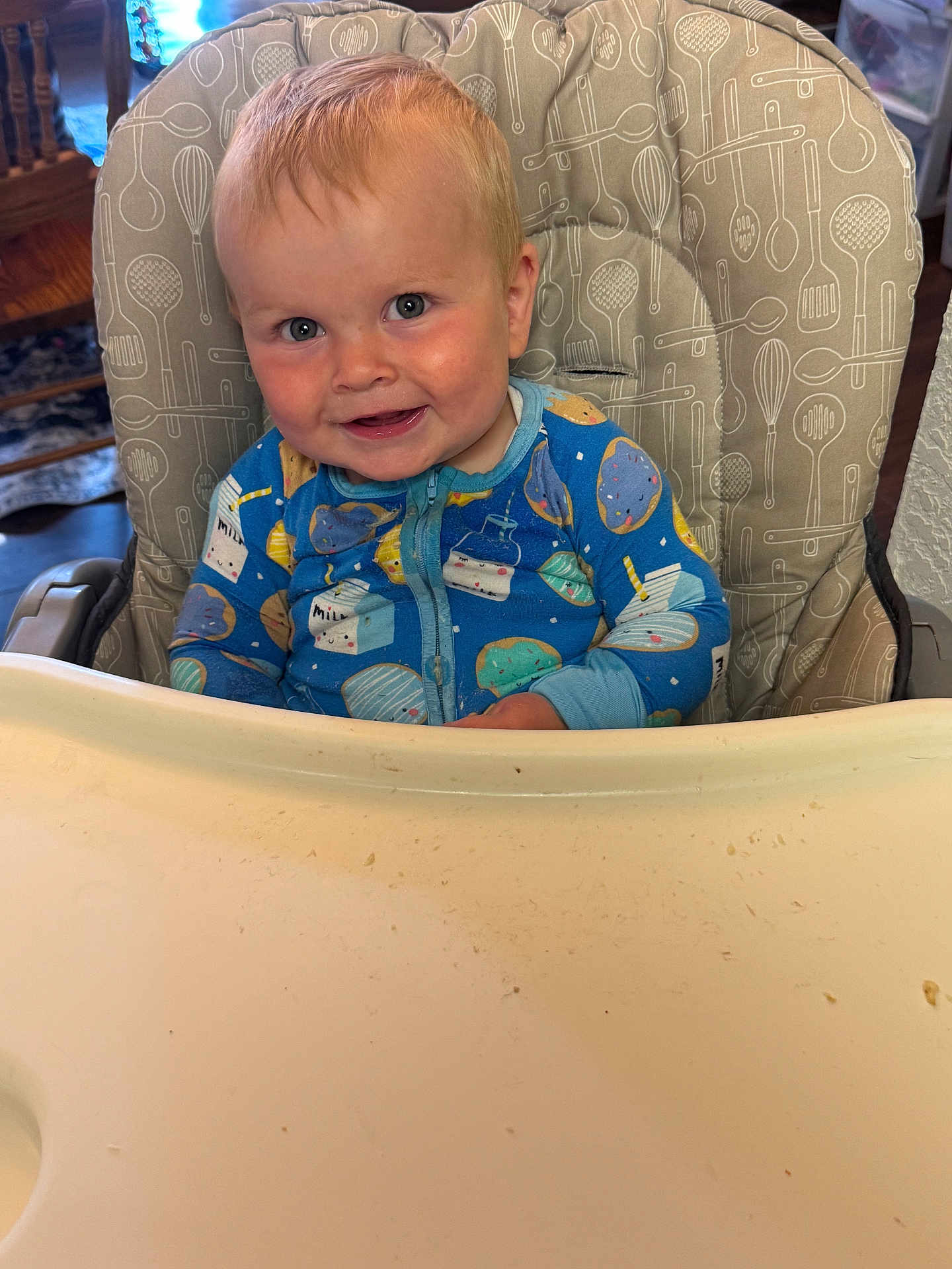 Eli is registered to the contest to win money with this photo: baby, child, high_chair, onesie, blue_clothing, smiling, face, indoor, messy_tray, milk_carton_pattern, donut_pattern, cute, infant, happy, person, furniture, wooden_chair, table, home, portrait