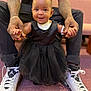 baby, child, smiling, black_dress, pink_bow, tattooed_arms, white_sneakers, holding_hands, indoor, carpet, couch, person, cute, happy, portrait, support, fashion, footwear, seated_adult, closeup