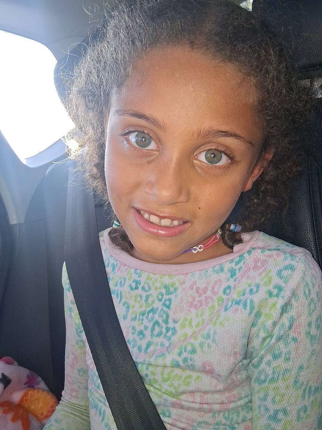 beaded_necklace, car_seat, casual_clothing, child, curly_hair, cute, face, happy, indoors, interior, patterned_shirt, person, portrait, seat, seatbelt, smile, sunlight, travel, window, young