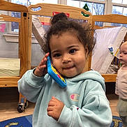 Gracie joined the competition — help win amazing prizes! toddler, child, toy_phone, hoodie, indoor, daycare, carpet, wooden_furniture, baby, play, cute, person, floor, window, clothing, face, expression, activity, young_child, casual