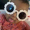 apparel, baby, child, closeup, cute, expression, face, fashion, flower_shaped_glasses, funny, headwear, indoor, infant, person, pink_clothing, playful, portrait, smile, sunglasses, tongue_out