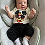 baby, infant, mickey_mouse, shirt, black_pants, white_socks, cushion, seat, soft_fabric, smiling, face, child, indoors, comfortable, clothing, cute, young, person, relaxing, baby_seat