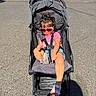 child, denim_shorts, face, hair, harness, heart_shaped_sunglasses, outdoor, parent, parked_cars, parking_lot, pavement, pink_shirt, portrait, sandals, seated, stroller, sunglasses, sunlight, toddler, topknot