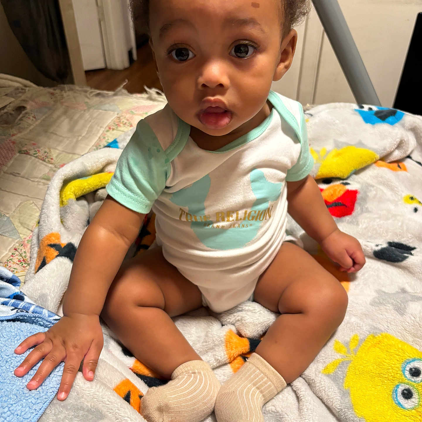 Aizhon joined the competition — help win amazing prizes! baby, child, indoor, blanket, socks, onesie, curly_hair, floor, wooden_floor, door, expression, cute, toddler, hand, face, seated, colorful, patterned_blanket, person, home