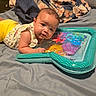 baby, infant, bed, blanket, pillow, toy, inflatable, water_mat, curious, lying_down, indoor, home, soft, colorful, child, person, face, clothing, yellow_pants, white_shirt