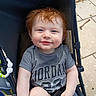 toddler, child, smile, red_hair, blue_eyes, stroller, tshirt, sitting, outdoor, pavement, happy, person, baby, casual_clothing, hand, face, portrait, young_child, cute, sunlight