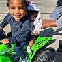 toddler, baby, toy_atv, green_vehicle, smiling_child, braided_hair, hooded_jacket, adult_hand, sunny_day, outdoor, street, sidewalk, shoes, socks, playtime, child, family, support, vehicle, happy