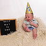 baby, infant, party_hat, celebration, milestone, signboard, text, fluffy_rug, smiling, happy, cute, sitting, clothing, shorts, shirt, studio, portrait, child, indoor, young