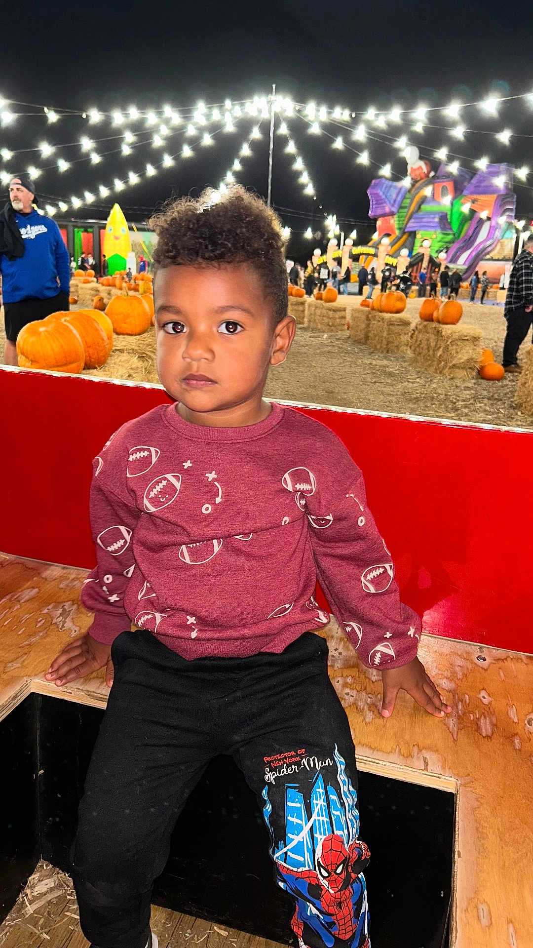 Jeffer is registered to the contest to win money with this photo: child, pumpkin_patch, night, string_lights, hay_bales, pumpkins, inflatable_slide, people, bench, clothing, football_pattern, spiderman, pants, wood, fall, festival, outdoor, casual, serious, event