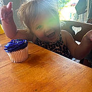 Ivory is registered to the contest to win money with this photo: toddler, child, cupcake, purple_frosting, wooden_table, smile, happy, indoor, sunlight, window, chair, celebration, food, birthday, person, clothing, hair, face, hands, home