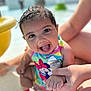 Rosalia is registered to the contest to win money with this photo: baby, child, smiling, wet_hair, swimsuit, colorful_clothing, hand_holding, water, pool, sunlight, happy, sitting, person, cute, summer, outdoor, playful, skin, face, toddler