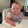 baby, child, high_chair, bib, smile, indoor, face, hand, furniture, window, fan, clothing, person, home, expression, food, hair, seat, room, table