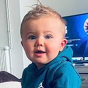 Tao a rejoint le concours — aidez-le/la à gagner de superbes lots ! baby, blue_eyes, casual, child, clothing, curly_hair, cute, face, furniture, home, hoodie, indoor, living_room, person, portrait, seated, smiling, television, toddler, young_child