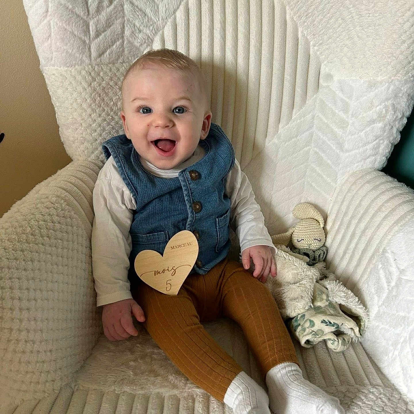 Marceau S a rejoint le concours — aidez-le/la à gagner de superbes lots ! armchair, baby, blanket, blue_eyes, brown_leggings, chubby_cheeks, cozy, denim_vest, happy, indoor, infant, long_sleeve, milestone_card, plush_toy, portrait, seated, smiling, textured_fabric, white_socks, wooden_heart