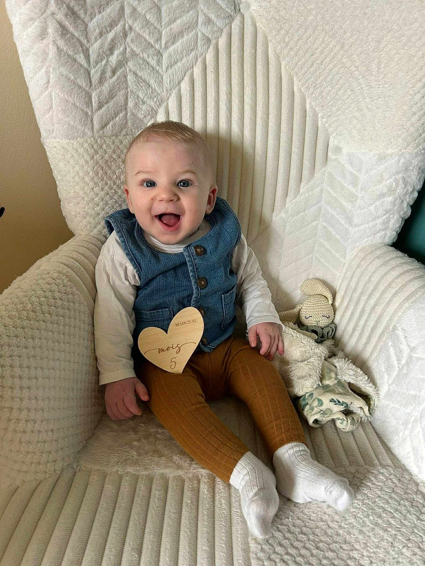 Marceau S a rejoint le concours — aidez-le/la à gagner de superbes lots ! baby, infant, smiling, seated, milestone_card, wooden_heart, denim_vest, long_sleeve, brown_leggings, white_socks, plush_toy, blanket, armchair, textured_fabric, cozy, portrait, happy, blue_eyes, chubby_cheeks, indoor