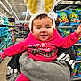 adult_hand, aisle, baby, bunny_ears, child, colorful, happy, headband, holding, infant, person, pink_hoodie, portrait, princess, retail, shelves, shopping, smiling, supermarket, toys