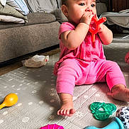 Raven joined the competition — help win amazing prizes! baby, infant, child, teething_toy, toy, rattle, pink_clothing, sitting, play_mat, living_room, sofa, barefoot, biting, colorful_toys, crawling_mat, curious_expression, face, hands, floor, portrait