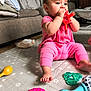 baby, infant, child, teething_toy, toy, rattle, pink_clothing, sitting, play_mat, living_room, sofa, barefoot, biting, colorful_toys, crawling_mat, curious_expression, face, hands, floor, portrait