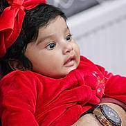 Anette is registered to the contest to win money with this photo: baby, child, infant, red_clothing, bow, face, portrait, cute, gaze, adult_hand, watch, closeup, soft_texture, indoor, warm_colors, curly_hair, expression, holding, toddler, person