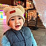 toddler, child, hat, knit_hat, pom_poms, vest, blue_jacket, smile, happy, face, outdoor, evening, lights, festive, urban, pavement, portrait, warm_clothing, cute, winter