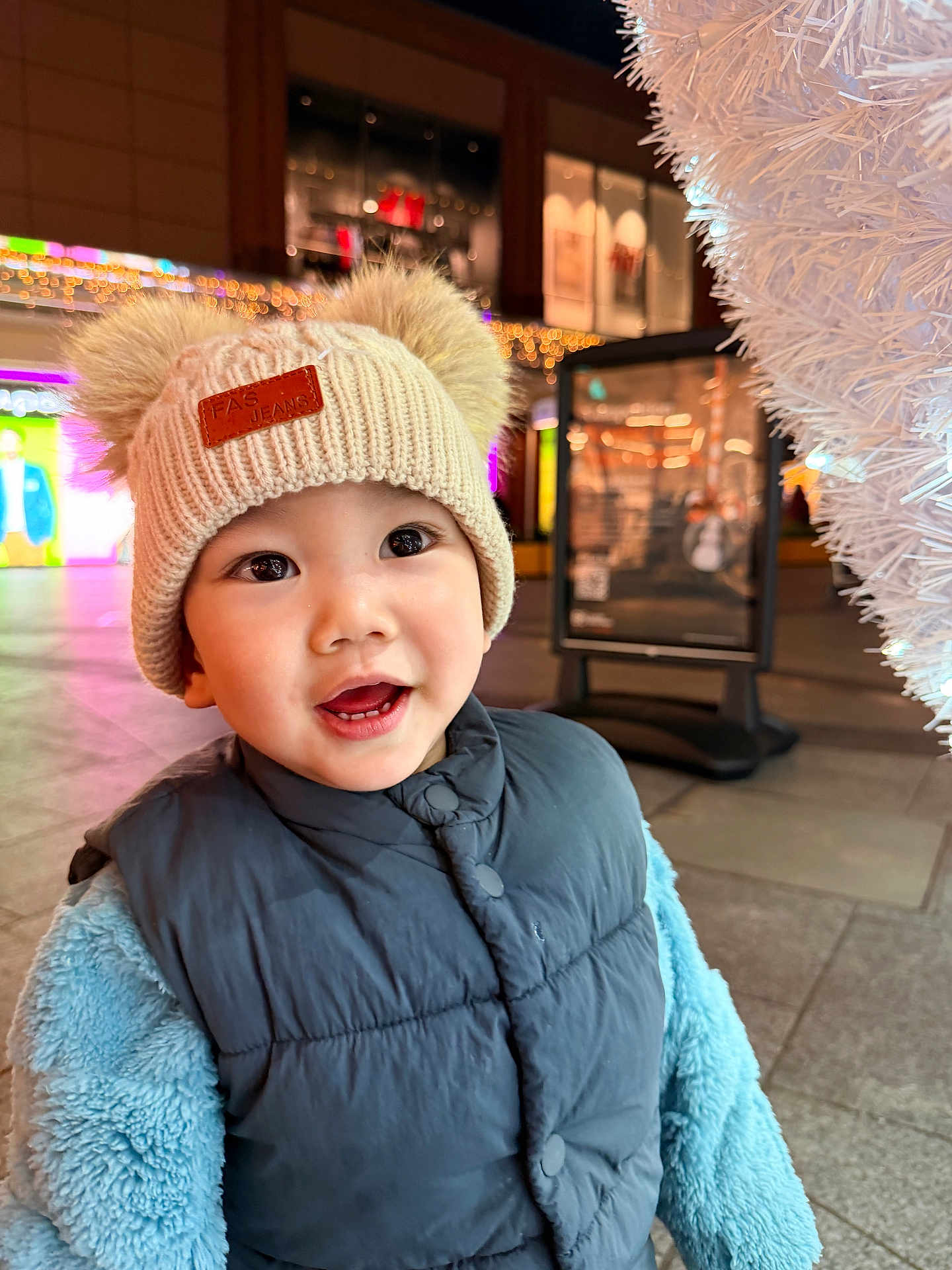Julian is registered to the contest to win money with this photo: toddler, child, hat, knit_hat, pom_poms, vest, blue_jacket, smile, happy, face, outdoor, evening, lights, festive, urban, pavement, portrait, warm_clothing, cute, winter