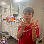 child, boy, red_shirt, peace_sign, bathroom, mirror, cabinet, floor, lighting, expression, clutter, clothing, person, indoor, hand_gesture, reflection, countertop, plastic_container, wood_floor, casual