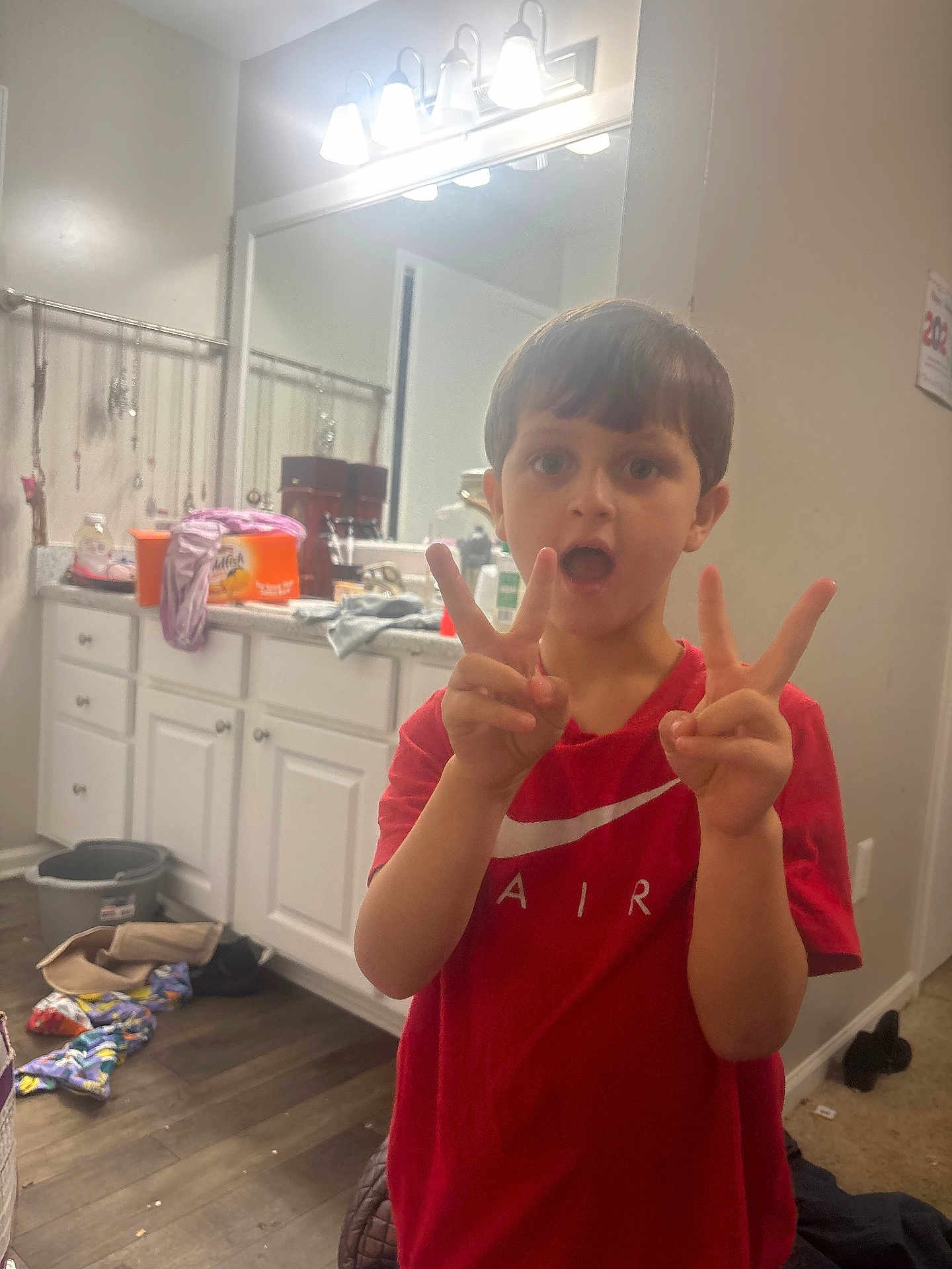 Ty Sears is registered to the contest to win money with this photo: child, boy, red_shirt, peace_sign, bathroom, mirror, cabinet, floor, lighting, expression, clutter, clothing, person, indoor, hand_gesture, reflection, countertop, plastic_container, wood_floor, casual