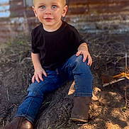 Austin is registered to the contest to win money with this photo: toddler, child, boy, blue_eyes, black_tshirt, jeans, boots, sitting, outdoor, dirt, top_knot, curious, portrait, daylight, rustic, wooden_wall, casual_clothing, young, cute, nature