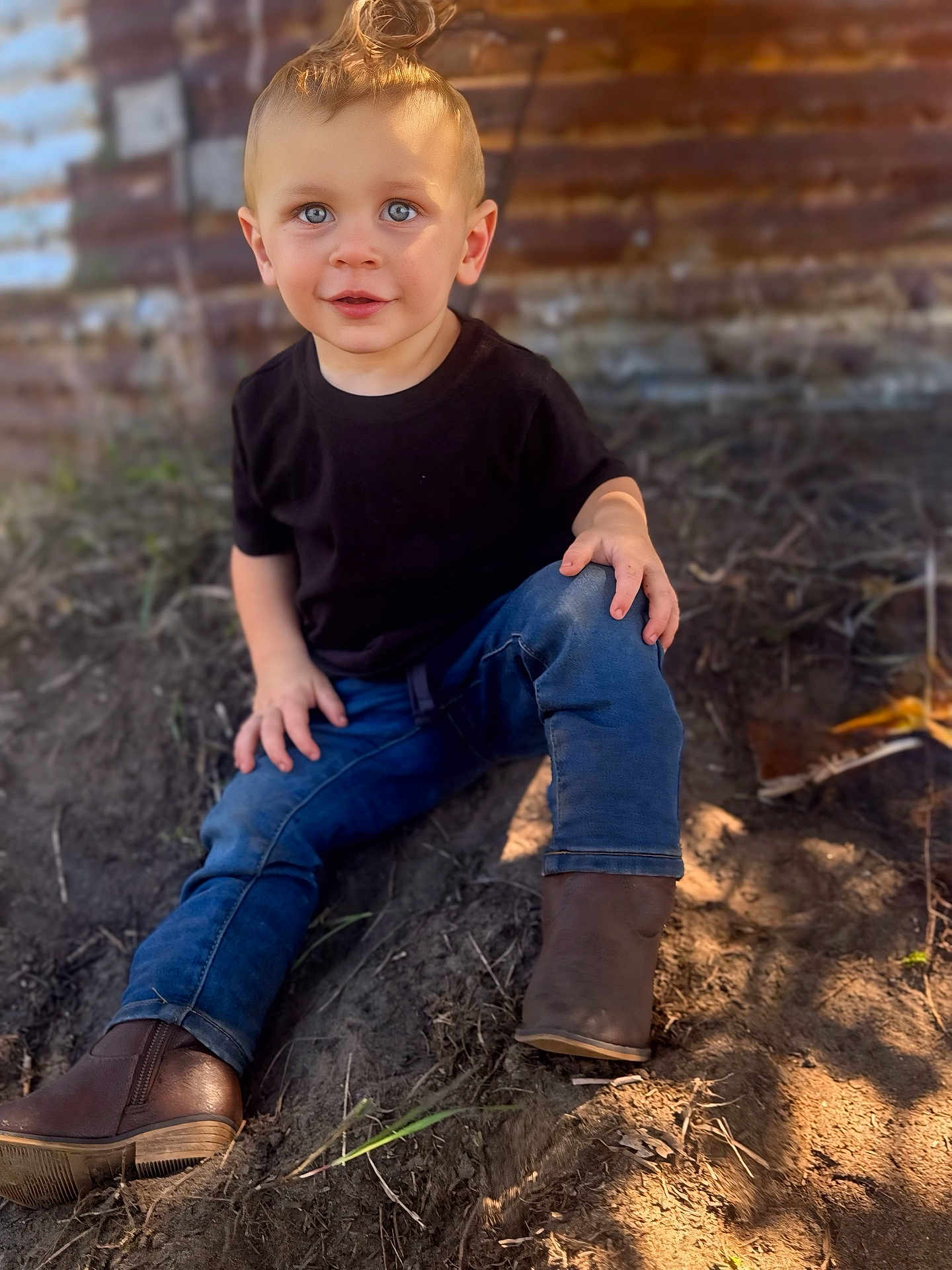 Austin is registered to the contest to win money with this photo: toddler, child, boy, blue_eyes, black_tshirt, jeans, boots, sitting, outdoor, dirt, top_knot, curious, portrait, daylight, rustic, wooden_wall, casual_clothing, young, cute, nature
