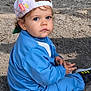 backward_cap, blue_eyes, blue_tracksuit, cap, casual_clothing, child, curly_hair, cute, daylight, gravel, nature, outdoor, person, portrait, serious_face, shoes, sitting, small_hands, toddler, young_child