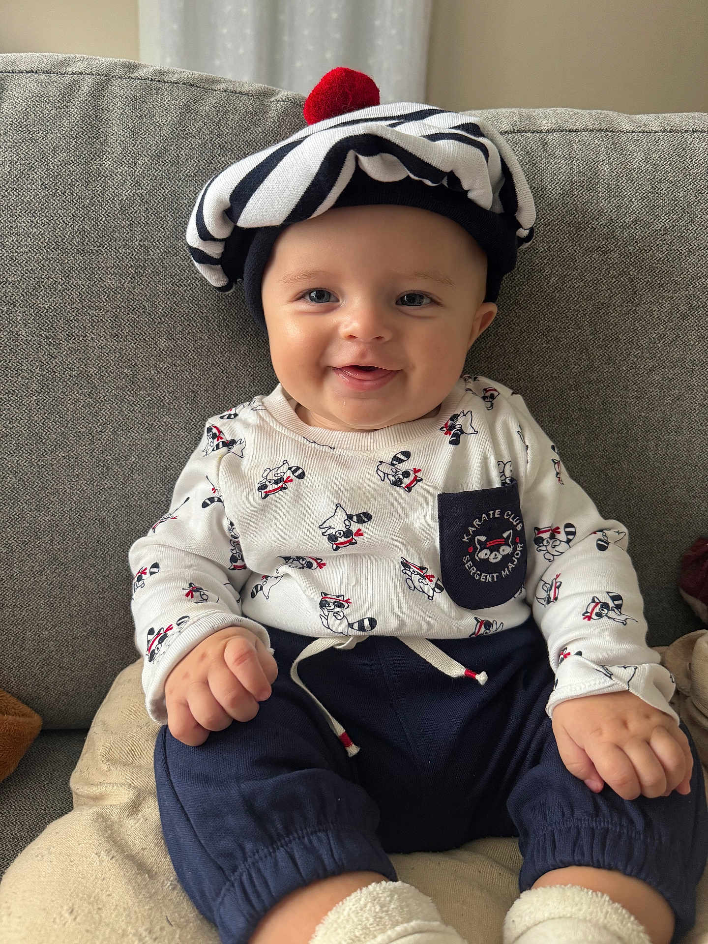 Léon participe au concours pour gagner de l'argent avec cette photo : baby, child, smiling, hat, pom_pom, striped_hat, couch, clothing, raccoon_pattern, long_sleeve_shirt, pants, socks, indoors, person, cute, happy, seated, face, infant, portrait