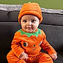 baby, child, pumpkin_costume, orange_clothing, hat, cute, smiling, sitting, indoor, couch, green_wall, face, portrait, infant, cozy, seasonal, festive, happy, person, costume