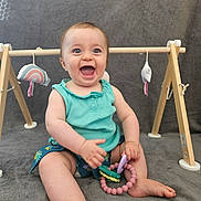 Margot a rejoint le concours — aidez-le/la à gagner de superbes lots ! baby, child, smiling, teething_toy, play_gym, wooden_toy, blue_top, shorts, barefoot, sitting, happy, indoor, gray_blanket, cute, fun, infant, person, toy, playtime, portrait