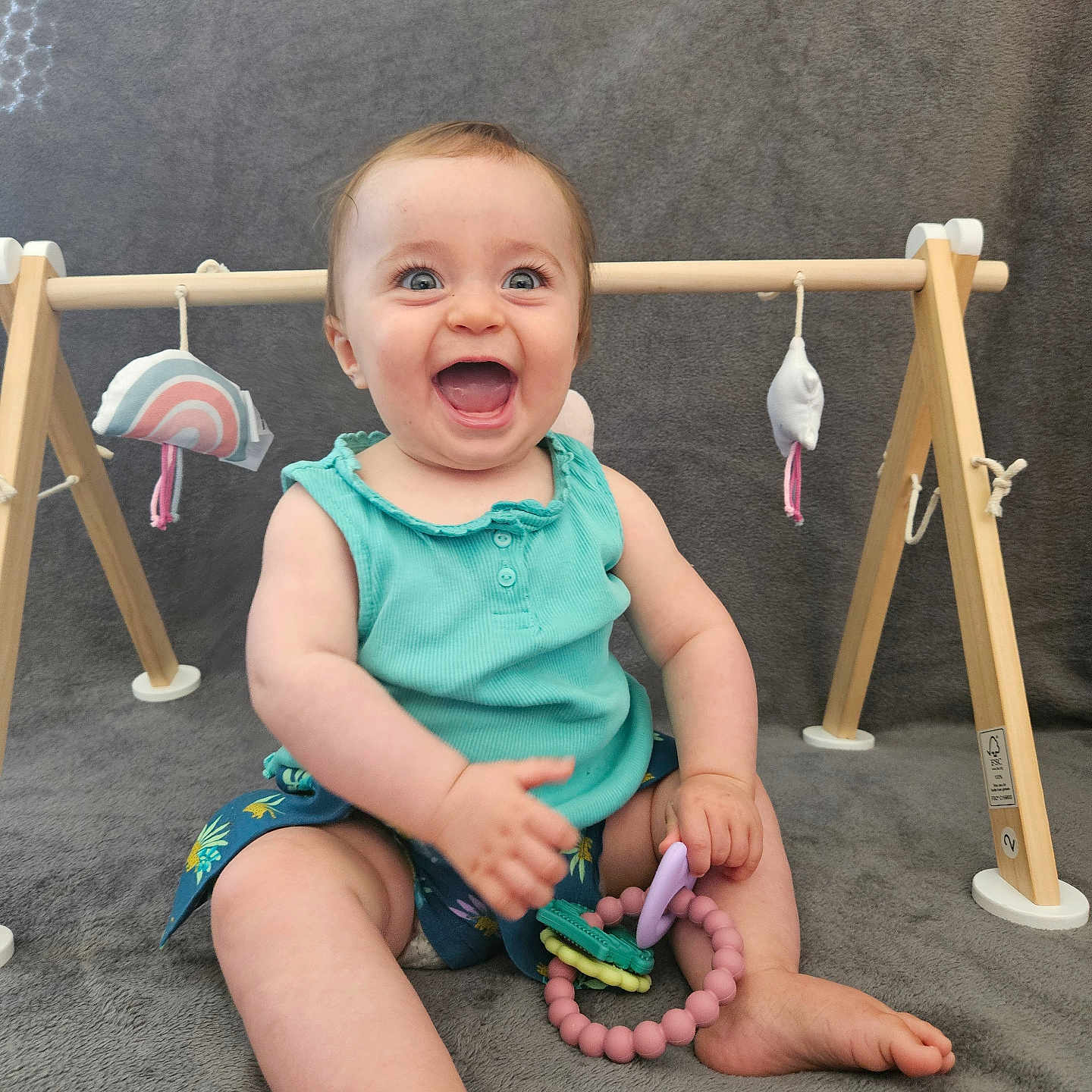 Margot a rejoint le concours — aidez-le/la à gagner de superbes lots ! baby, barefoot, blue_top, child, cute, fun, gray_blanket, happy, indoor, infant, person, play_gym, playtime, portrait, shorts, sitting, smiling, teething_toy, toy, wooden_toy