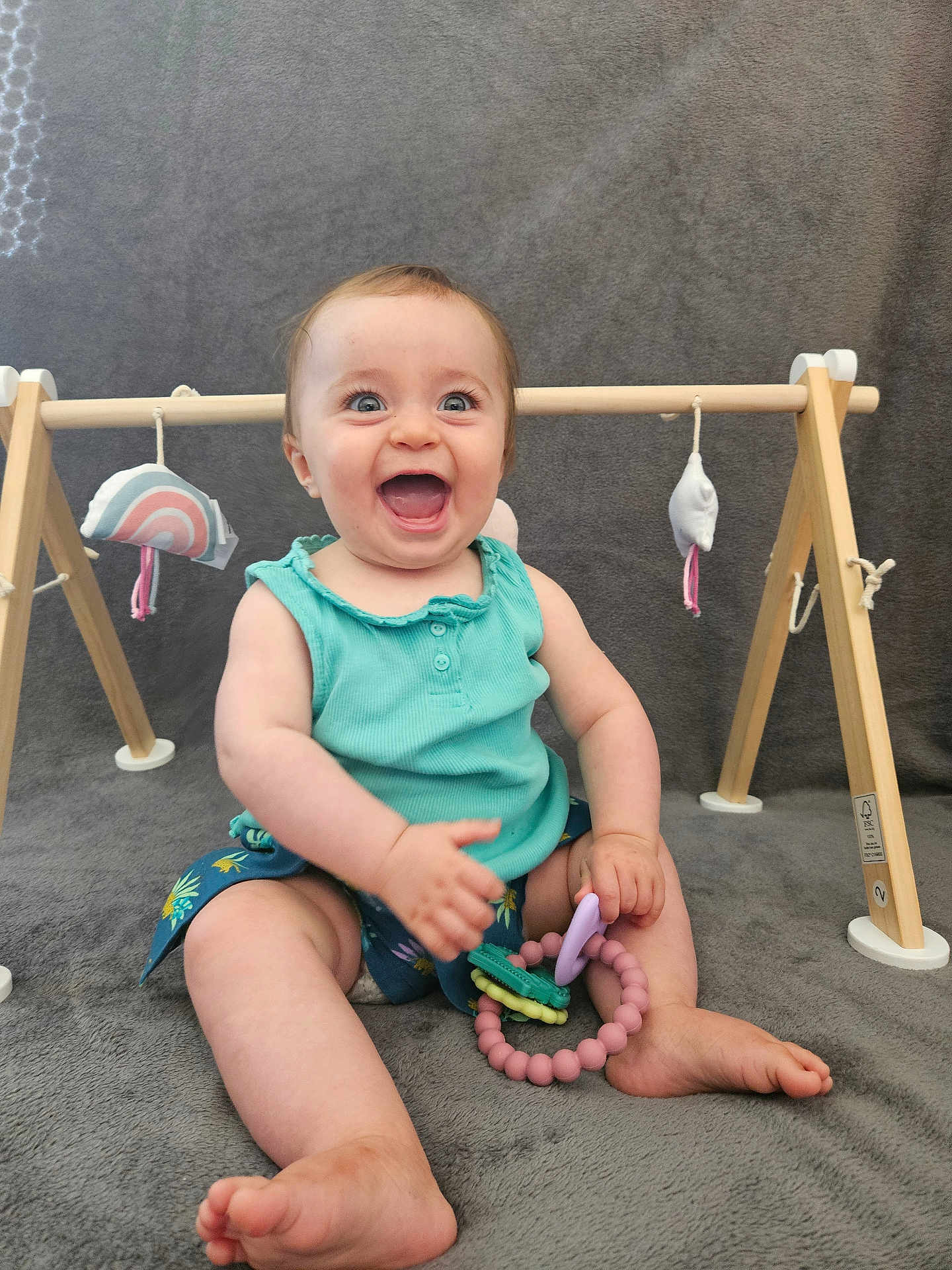 Margot a rejoint le concours — aidez-le/la à gagner de superbes lots ! baby, child, smiling, teething_toy, play_gym, wooden_toy, blue_top, shorts, barefoot, sitting, happy, indoor, gray_blanket, cute, fun, infant, person, toy, playtime, portrait