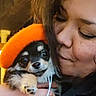 person, dog, puppy, hat, orange_hat, close_up, indoor, affection, cuddle, warm_lighting, black_clothing, bracelet, cute, pet, head, face, hand, fur, small_dog, love
