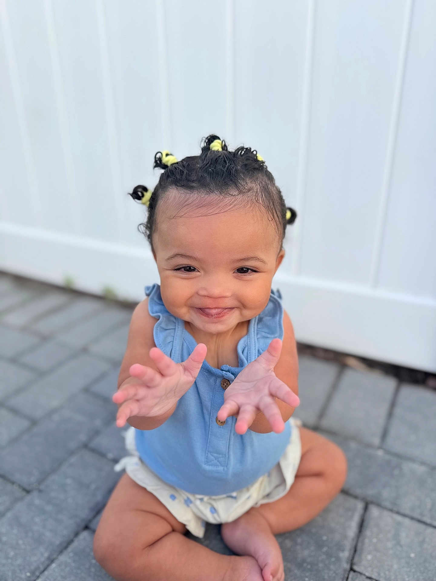 Leilani joined the competition — help win amazing prizes! toddler, child, smiling, sitting, pavement, blue_top, hair_buns, yellow_hair_ties, happy, cute, outdoor, baby, skin, hands, face, portrait, playful, young_child, expression, fence