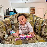Leilani is registered to the contest to win money with this photo: baby, child, smiling, toy, armchair, patterned_furniture, indoor, living_room, ceiling_fan, window_blinds, colorful, happy, person, seated, curly_hair, infant, playful, home, cozy, casual