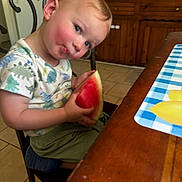 Seth joined the competition — help win amazing prizes! toddler, child, watermelon, food, chair, kitchen, table, placemat, wooden_cabinet, curious, rosy_cheeks, short_sleeve_shirt, dinosaur_print, green_shorts, indoor, person, face, sitting, cute, snack