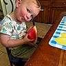 toddler, child, watermelon, food, chair, kitchen, table, placemat, wooden_cabinet, curious, rosy_cheeks, short_sleeve_shirt, dinosaur_print, green_shorts, indoor, person, face, sitting, cute, snack