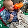 baby, child, toddler, sippy_cup, orange_bottle, blanket, pillow, cushion, drinking, sitting, face, eyes, hand, blue_shirt, portrait, outdoor, patio_heater, metal_tank, cozy, soft_texture