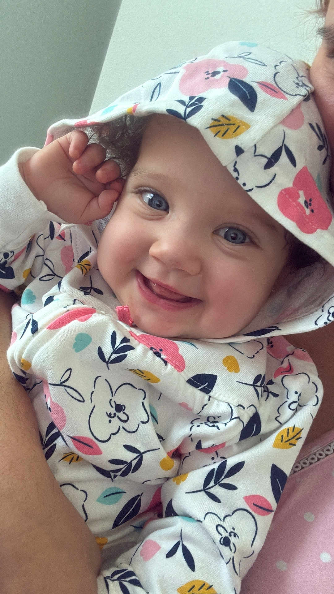 Samantha is registered to the contest to win money with this photo: baby, child, smile, hoodie, patterned_clothing, blue_eyes, curly_hair, cute, happy, portrait, indoor, closeup, infant, person, face, hand, floral_print, soft_lighting, childhood, warmth