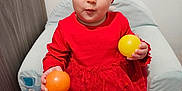 Yzïa a rejoint le concours — aidez-le/la à gagner de superbes lots ! toddler, child, red_dress, headband, ball, yellow_ball, orange_ball, pink_ball, cushioned_chair, play_mat, indoor, curious_expression, blue_eyes, sitting, soft_furniture, floor_mat, baby, playful, cute, portrait