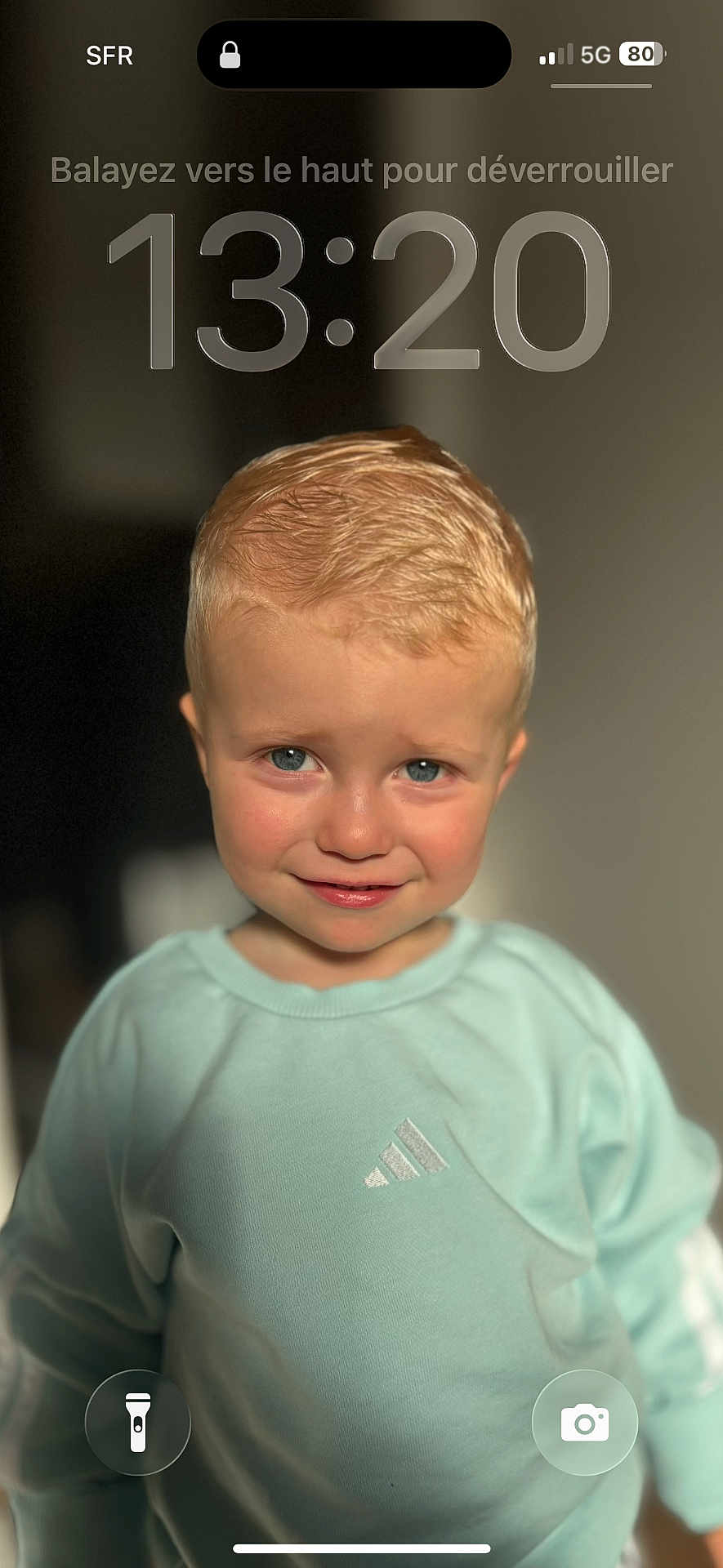 Jules participe au concours pour gagner de l'argent avec cette photo : child, toddler, boy, blond_hair, blue_eyes, face, portrait, close_up, smile, sweatshirt, mint_color, indoor, soft_light, bokeh, lock_screen, time_overlay, camera_icon, flashlight_icon, skin, hair
