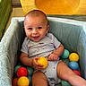 baby, bright_colors, child, colorful_balls, cute, fabric, face, hand, happy, indoor, infant, leg, onesie, playpen, playtime, seated, skin, smiling, texture, toy